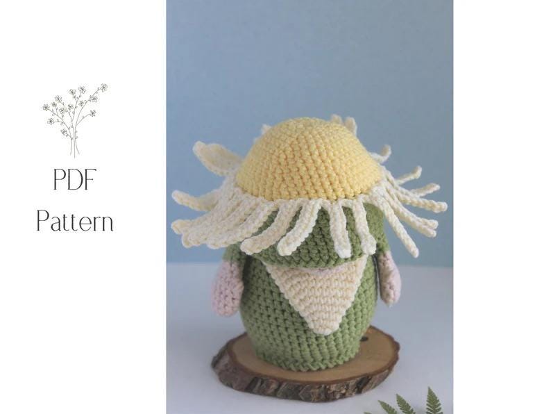 May include: A crocheted gnome doll with a yellow and white flower hat, green body, and a cream-colored scarf. The doll is sitting on a wooden slice. The words "PDF Pattern" are in the upper left corner.