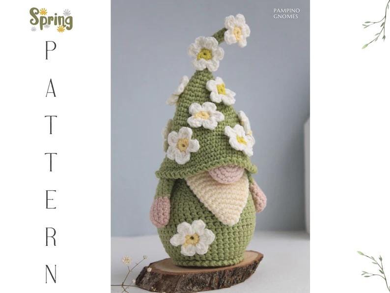 May include: A handmade crochet gnome with a green body and hat, adorned with white and yellow flower accents. The gnome has a beige beard, pink hands, and is displayed on a wooden slice. The image includes the text "Spring Pattern".