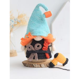 May include: A crocheted gnome doll wearing a blue hat with an orange brim, a black and brown vest with orange accents, and holding a yellow crocheted hairdryer. The gnome is sitting on a wooden base.