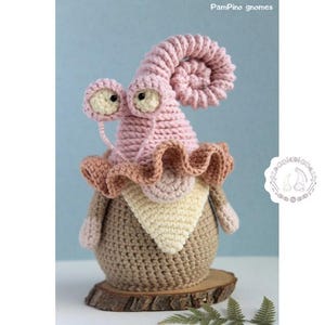 May include: A handcrafted crochet gnome featuring a snail-shaped hat and a frilled collar. The gnome is predominantly pink and brown, with a cream-coloured bib. The text "PamPino gnomes" is visible in the upper right corner.