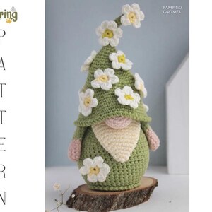 May include: A handmade crochet gnome with a green hat adorned with white and yellow daisy flowers. The gnome has a pink nose, a cream-colored beard, and pink hands. It is displayed on a wooden slice. The image includes the text "Spring Pattern".