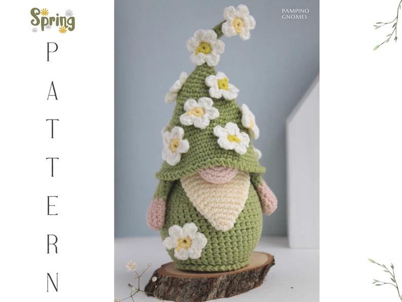 May include: A handmade crochet gnome with a green hat adorned with white and yellow daisy flowers. The gnome has a pink nose, a cream-colored beard, and pink hands. It is displayed on a wooden slice. The image includes the text "Spring Pattern".
