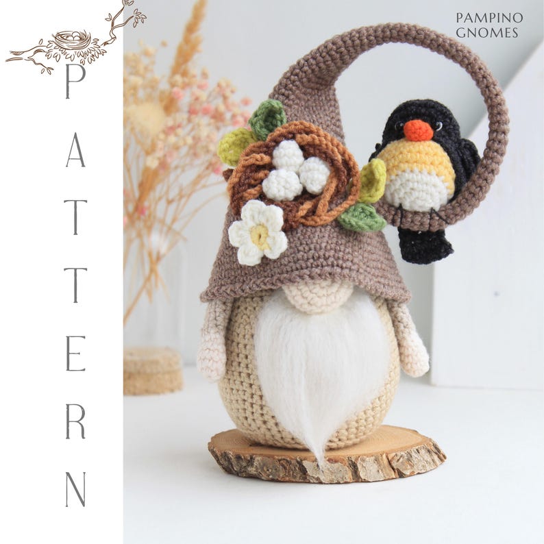 Puede incluir: Una figura de gnomo hecha a mano con un gorro de ganchillo marr&oacute;n, una barba blanca y un p&aacute;jaro posado en un lazo. El gnomo est&aacute; adornado con un nido, flores y hojas. La base es una rodaja de madera. El texto "PAMPINO GNOMES" es visible.