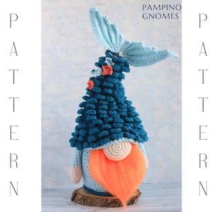 Puede incluir: Un gnomo de ganchillo hecho a mano con un gorro y cola azules, barba naranja y cara color crema. El gnomo está decorado con pequeñas flores de color coral. El texto "PAMPINO GNOMES" y "PATTERN" son visibles.