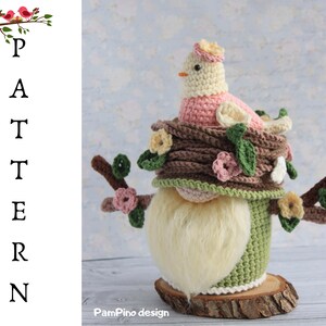 May include: A handcrafted crochet gnome with a bird on a nest. The gnome features a long, fluffy white beard, a green body, and a brown hat decorated with flowers. The bird is pink and cream, with a small yellow beak. The gnome is on a wooden base.