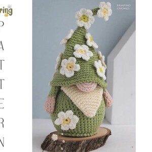 May include: A handmade crochet gnome with a green hat and body, adorned with white and yellow daisy flowers. The gnome has a beige beard and pink hands and nose. The gnome is displayed on a wooden slice. Text on the image says "Spring Pattern".