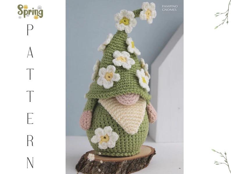 May include: A handmade crochet gnome with a green hat and body, adorned with white and yellow daisy flowers. The gnome has a beige beard and pink hands and nose. The gnome is displayed on a wooden slice. Text on the image says "Spring Pattern".