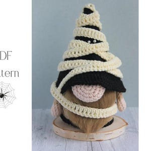Op de afbeelding: Een handgemaakte Halloween-kabouter verkleed als mummie. De kabouter heeft een zwarte hoed, een bruine baard en een roze neus. De mummiewikkelingen zijn van crèmekleurig garen. De kabouter staat op een houten voet. De tekst "PDF Pattern" staat in de afbeelding.