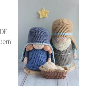 May include: Crocheted nativity scene featuring two figures in blue and tan, with a small basket and a yellow star. The figures have rounded bodies and are wearing hats and robes. The text "PDF Pattern" is visible.