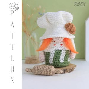 Puede incluir: Un gnomo de ganchillo hecho a mano con un gorro de chef blanco y un delantal a rayas verdes y blancas. El gnomo tiene pelo naranja y un pequeño pastel marrón en su sombrero. Un pequeño rodillo de ganchillo beige está delante del gnomo.