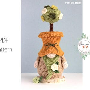 May include: A handmade crochet gnome with an orange hat and a green beard, holding a small tree with green and white flowers. The gnome is tan and sits on a wooden base. The text "PDF Pattern" is on the left.