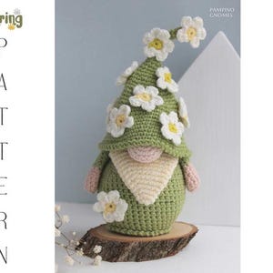 May include: A handmade crochet gnome with a green hat adorned with white and yellow flower accents. The gnome has a beige beard and pink hands and nose. The gnome is displayed on a wooden slice. Text on the left says "Spring Pattern".
