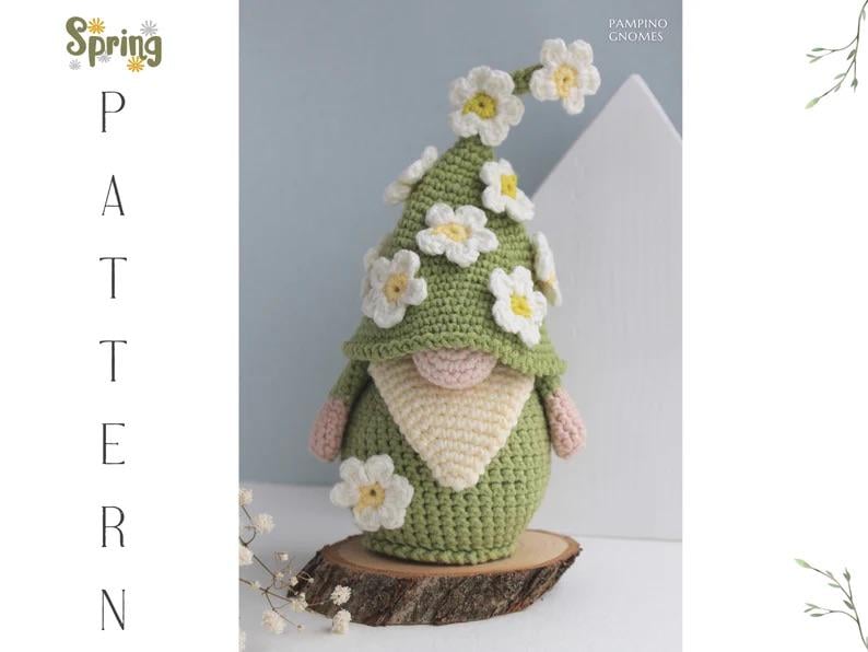 May include: A handmade crochet gnome with a green hat adorned with white and yellow flower accents. The gnome has a beige beard and pink hands and nose. The gnome is displayed on a wooden slice. Text on the left says "Spring Pattern".