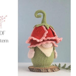 May include: A crocheted gnome figurine with a red and pink flower-shaped hat and a green body. The gnome has a cream-colored beard and is displayed on a wooden slice. The image also includes the text "PDF Pattern".
