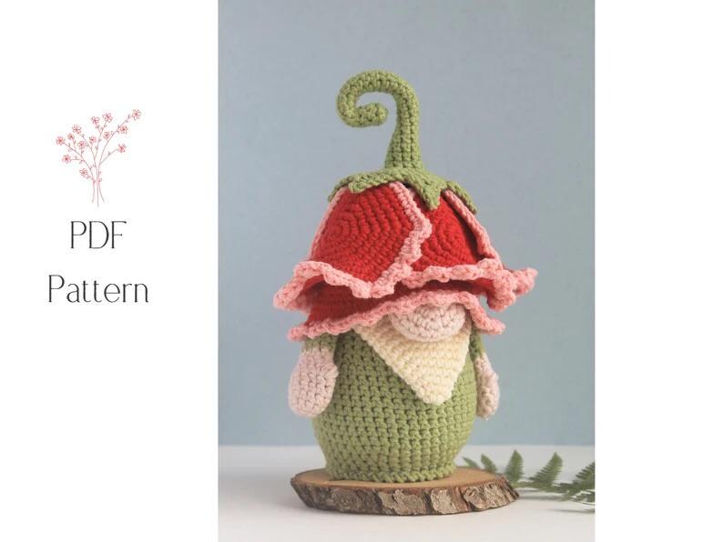 May include: A crocheted gnome figurine with a red and pink flower-shaped hat and a green body. The gnome has a cream-colored beard and is displayed on a wooden slice. The image also includes the text "PDF Pattern".