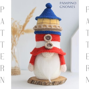 Può includere: Uno gnomo all'uncinetto fatto a mano a forma di faro, con una combinazione di colori blu, giallo, rosso e bianco. Lo gnomo ha una lunga barba bianca ed è esposto su una fetta di legno. I testi "PAMPINO GNOMES" e "PATTERN" sono visibili.