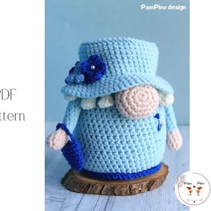 May include: A light blue crochet gnome with a hat, flower accents, and a small purse. The gnome has a pink nose and is sitting on a wooden base. The image includes the text "PDF Pattern".