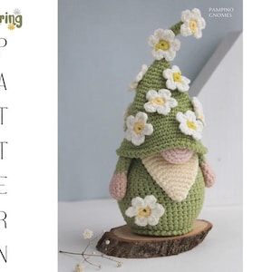 May include: A handmade crochet gnome with a green hat adorned with white and yellow flowers. The gnome has a pink nose and arms, a cream-colored beard, and is displayed on a wooden slice. The image includes the text "Spring Pattern".