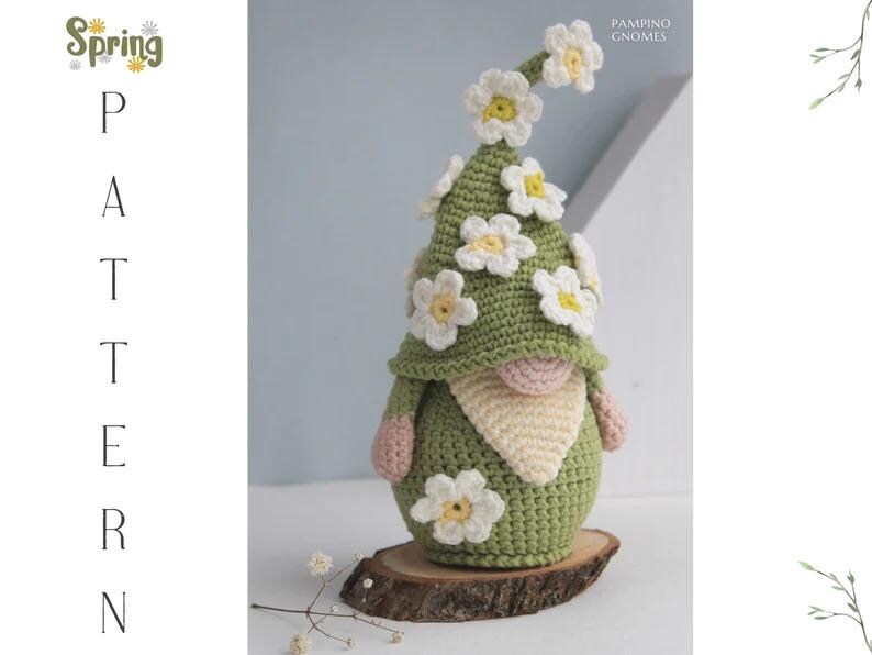 May include: A handmade crochet gnome with a green hat adorned with white and yellow flowers. The gnome has a pink nose and arms, a cream-colored beard, and is displayed on a wooden slice. The image includes the text "Spring Pattern".