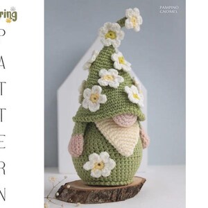 May include: A handmade crochet gnome with a green hat adorned with white and yellow daisy flowers. The gnome has a beige beard, pink hands, and is displayed on a wooden slice. Text on the left reads "Spring Pattern".