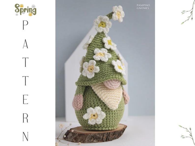 May include: A handmade crochet gnome with a green hat adorned with white and yellow daisy flowers. The gnome has a beige beard, pink hands, and is displayed on a wooden slice. Text on the left reads "Spring Pattern".