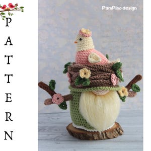 May include: A handmade crochet gnome featuring a bird in a nest design. The gnome has a long white beard, a green body, and a brown nest with a pink bird. The design includes floral accents and is set on a wooden base.