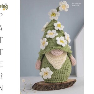 May include: A handmade crochet gnome with a green hat adorned with white and yellow flowers. The gnome has a beige beard, pink arms, and is displayed on a wooden base. The image includes the text "Spring Pattern".