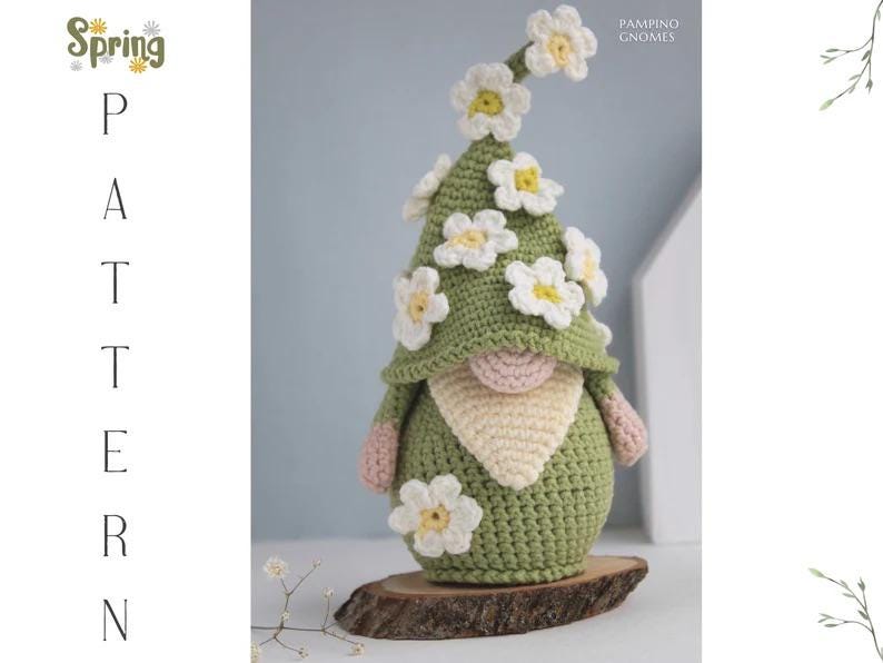 May include: A handmade crochet gnome with a green hat adorned with white and yellow flowers. The gnome has a beige beard, pink arms, and is displayed on a wooden base. The image includes the text "Spring Pattern".