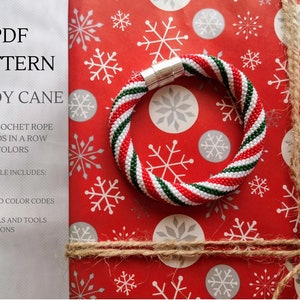 Puede incluir: Un brazalete de cuerda de crochet de cuentas rojo, blanco y verde en forma de bastón de caramelo. El brazalete está sobre un papel de envolver con estampado de copos de nieve rojos y blancos. El texto "PDF PATTERN CANDY CANE" es visible en la esquina superior izquierda de la imagen. El texto "BEAD CROCHET ROPE 17 BEADS IN A ROW 3 COLORS" es visible debajo del título. El texto "PDF FILE INCLUDES: CHARTS PHOTOS SEED BEAD COLOR CODES TIPS MATERIALS AND TOOLS SUGGESTIONS" es visible en la esquina inferior izquierda de la imagen.
