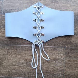 May include: A light blue corset-style belt with a central white cord lacing detail. The belt features silver-toned eyelets and a pointed top edge. The background is a wooden surface.