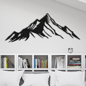 May include: Black metal mountain wall art. The artwork depicts a stylized mountain range with sharp peaks and valleys. The piece is mounted on a white wall above a gray sofa with white pillows and a bookshelf filled with books.