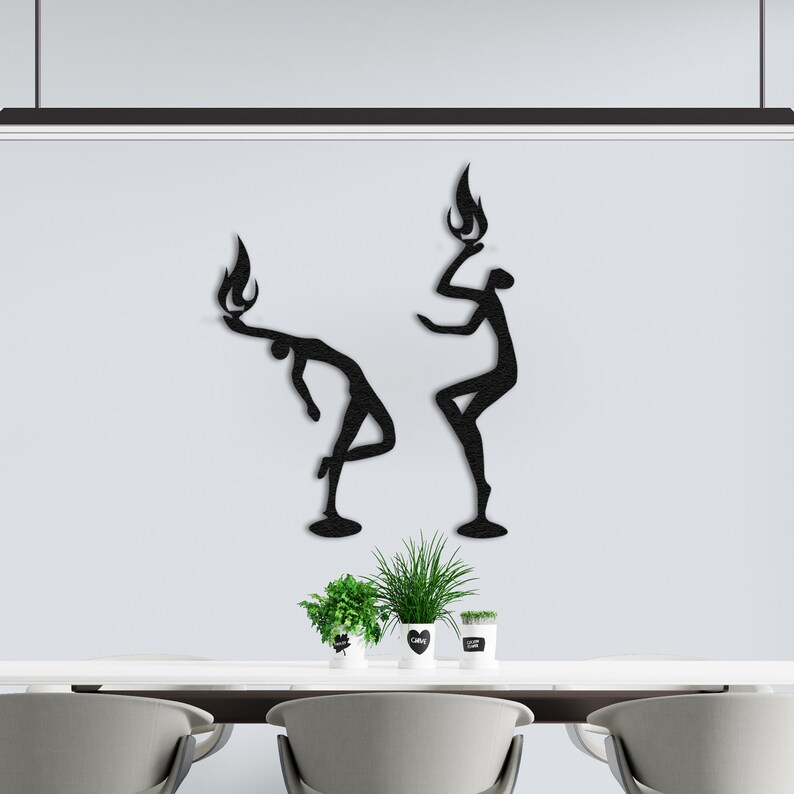 May include: Two black metal wall sculptures of stylized figures with flames on their heads. The figures are posed in a dynamic, abstract style. The sculptures are mounted on a white wall.