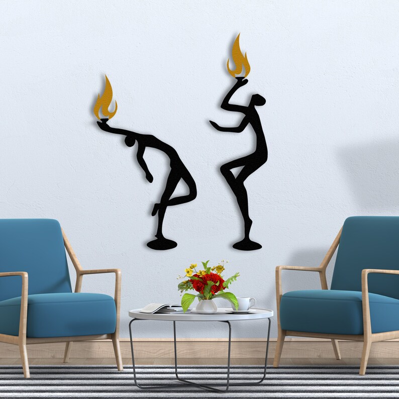May include: Two black metal wall sculptures of stylized figures holding flames above their heads. The figures are in a dancing pose. The flames are gold.