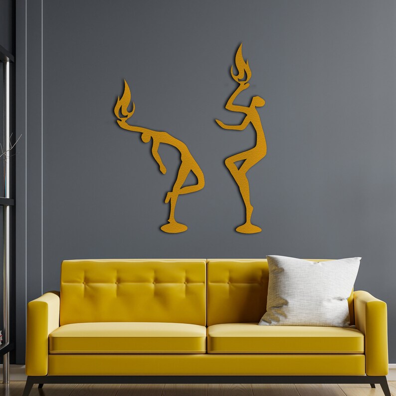 May include: Two gold-colored metal wall sculptures of stylized figures with flames on their heads. The figures are posed in a dynamic, abstract style. The sculptures are mounted on a gray wall above a yellow sofa.