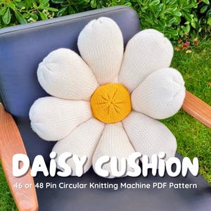 May include: A large, daisy-shaped cushion with white petals and a yellow center rests on a dark gray chair with wooden arms. The cushion is made of knitted fabric. Text on the image reads "DAISY CUSHION" and "46 or 48 Pin Circular Knitting Machine PDF Pattern."