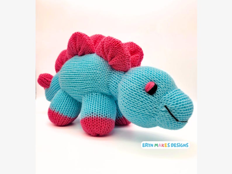 Light blue knitted Stegosaurus with pink spikes and feet. With the logo, Eryn Makes Designs.