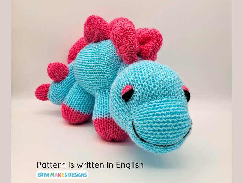 Light blue knitted Stegosaurus with pink spikes and feet. With the logo, Eryn Makes Designs, and the subtitle, Pattern is written in English.