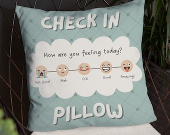 以下が含まれることがあります： 「CHECK IN」と「PILLOW」の文字が書かれた青と白の枕。枕は雲の形をしていて、5つの絵文字で表現された感情のスケールがあり、「良くない」から「素晴らしい！」までを表しています。