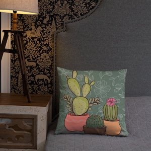 Puede incluir: Una almohada verde con un patr&oacute;n floral y un dise&ntilde;o de tres cactus en maceta. Los cactus son verdes, amarillos y rosas. Las macetas son naranja, rosa y marr&oacute;n.