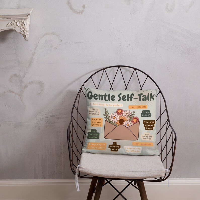 Puede incluir: Una almohada beige con un dise&ntilde;o floral y el texto "Gentle Self-Talk" en una fuente verde y marr&oacute;n. La almohada presenta un sobre marr&oacute;n con un borde blanco y un dise&ntilde;o floral. El sobre tiene varios cuadrados peque&ntilde;os con afirmaciones positivas escritas en texto negro.