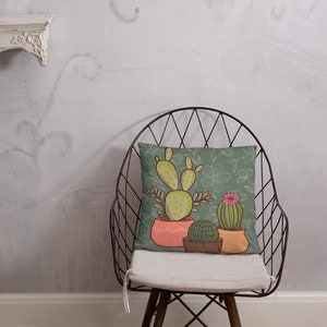 Puede incluir: Una almohada verde con un dise&ntilde;o de cactus. La almohada presenta tres cactus en macetas, con una maceta rosa, una maceta marr&oacute;n y una maceta amarilla. El fondo es un patr&oacute;n floral verde.