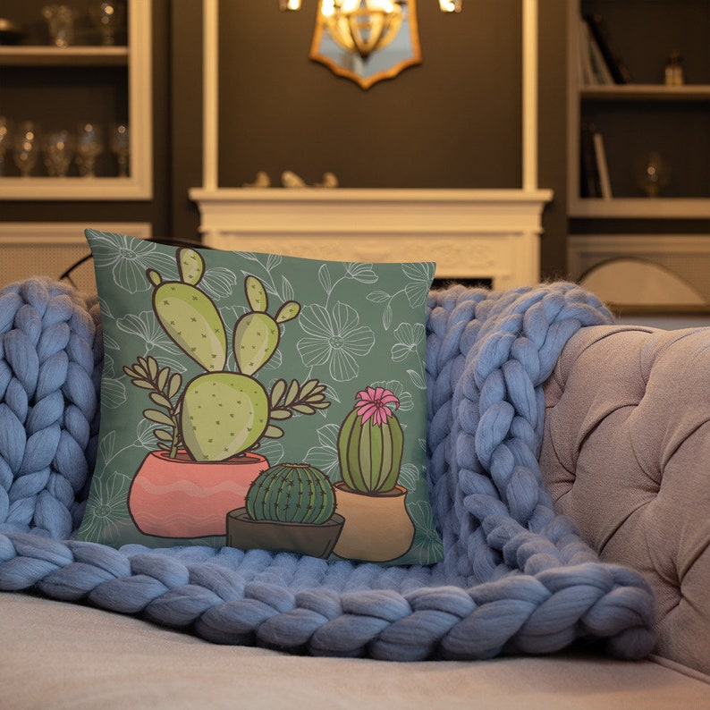 Puede incluir: Una almohada verde con un dise&ntilde;o de cactus. La almohada presenta tres cactus en macetas, uno con una flor rosa. El fondo es un patr&oacute;n floral verde.