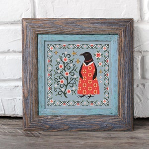 Puede incluir: Un bordado de punto de cruz enmarcado que presenta un pájaro negro con un vestido rojo con lunares blancos, de pie frente a un árbol floreciente con abejas volando a su alrededor. El fondo es azul claro con un borde blanco.