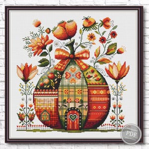 May include: A cross-stitch design of a pumpkin-shaped house with a heart-shaped door. The design incorporates floral and geometric patterns in orange, red, green, and yellow. The artwork is framed, and the measurements are in centimetres.