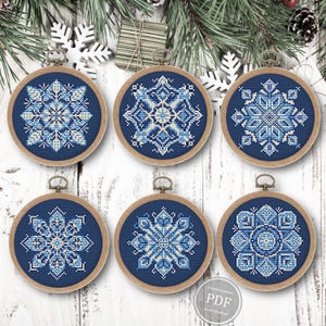 May include: Six circular wooden embroidery hoops containing cross-stitch patterns of intricate snowflakes in shades of blue, white, and cream on a dark blue fabric background. Each hoop features a unique geometric snowflake design.