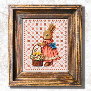 Könnte beinhalten: Kreuzstich-Bild eines Hasen in einem rosa Kleid, der einen Korb mit Blumen und Küken hält. Das gerahmte Kunstwerk hat einen braunen Holzrahmen. Der Hintergrund ist ein sich wiederholendes Muster aus orangefarbenen Blumen. Der Hase hat eine blaue Schleife.