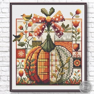 May include: Cross-stitch artwork depicting a pumpkin with a bow, birds, and floral details. The pumpkin is divided, with one side orange and the other white with a plaid design. The artwork is presented in a dark brown frame.