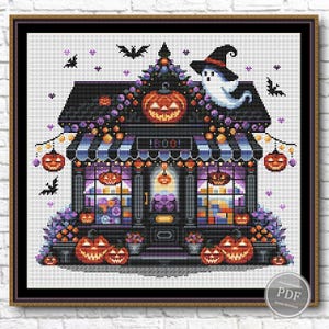 Puede incluir: Un patrón de punto de cruz de una tienda de Halloween espeluznante con un toldo a rayas blanco y negro, un fantasma con un sombrero de bruja y calabazas. La tienda está decorada con flores moradas y naranjas y luces. El letrero sobre la puerta dice "Boo!"
