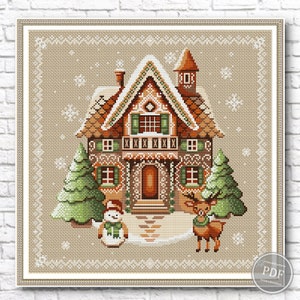 Gingerbread house cross stitch design with brown roof, green shutters, white trim, accompanied by snowman and two green trees on light brown background with white snowflakes.