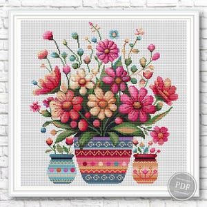 May include: Cross-stitch floral design featuring a vibrant bouquet of pink, red, and peach flowers in a decorative pot. Additional smaller vases with flowers are also present. The artwork is framed in a white border.